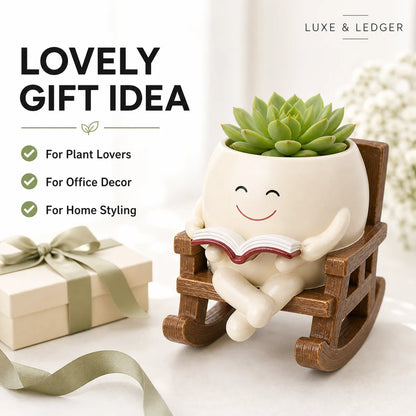Decorative planter with a smiling face reading a book, sitting on a small wooden rocking chair, with a gift box and ribbon in the background.