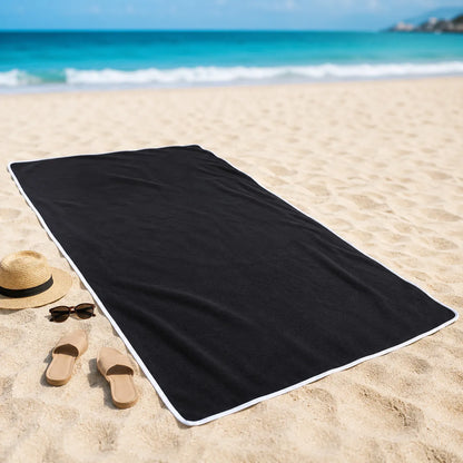 Black beach towel on a sandy beach with a hat, sandals, and sunglasses.