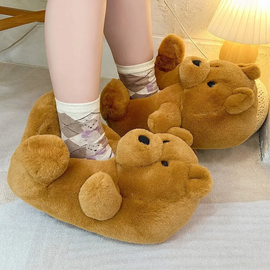 A person wearing plush teddy bear-shaped slippers in a cozy indoor setting. The slippers are made of soft brown faux fur and have embroidered black eyes and noses.