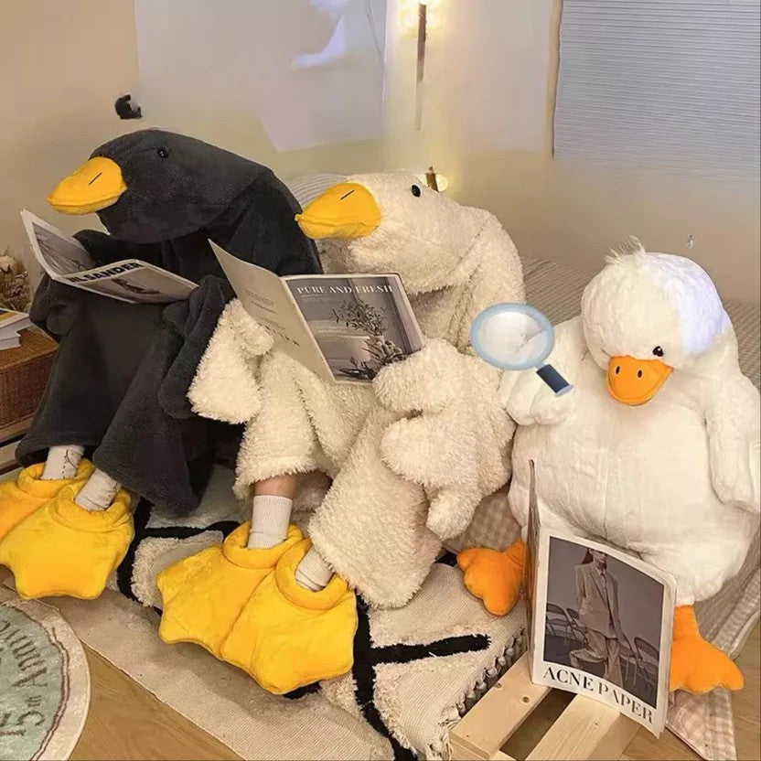 Three plush duck toys sitting on a couch with books, one wearing a black suit and yellow shoes, another in a white outfit with yellow shoes, and the third in a gray outfit with yellow shoes.