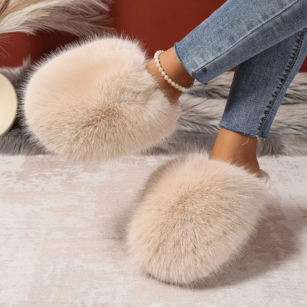 Fuzzy slippers with large pom-poms worn by a person in jeans on a soft surface.