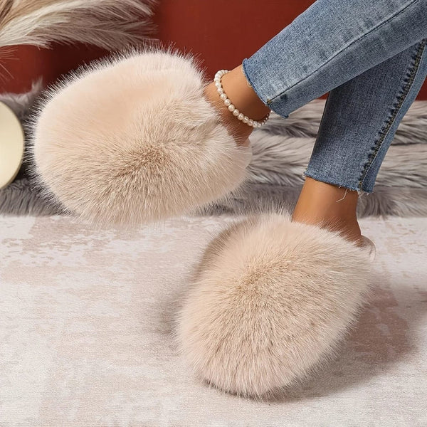 Fuzzy slippers with large pom-poms worn by a person in jeans on a soft surface.