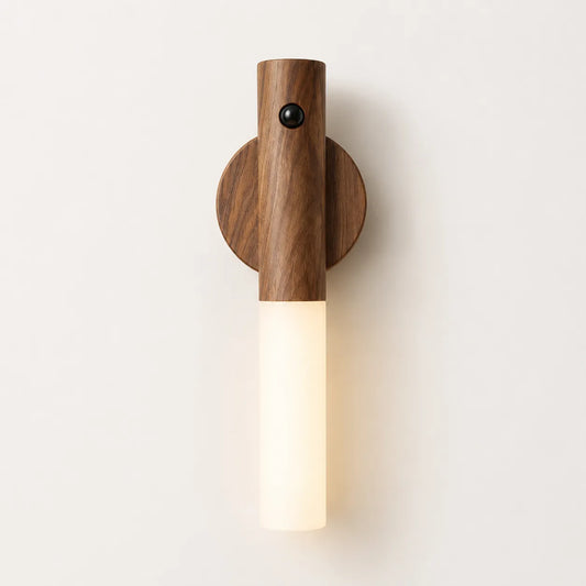 A modern wall-mounted light fixture with a dark wood cylindrical upper section, a circular wood backplate, and a glowing frosted white glass lower tube, shown against a plain off-white wall.