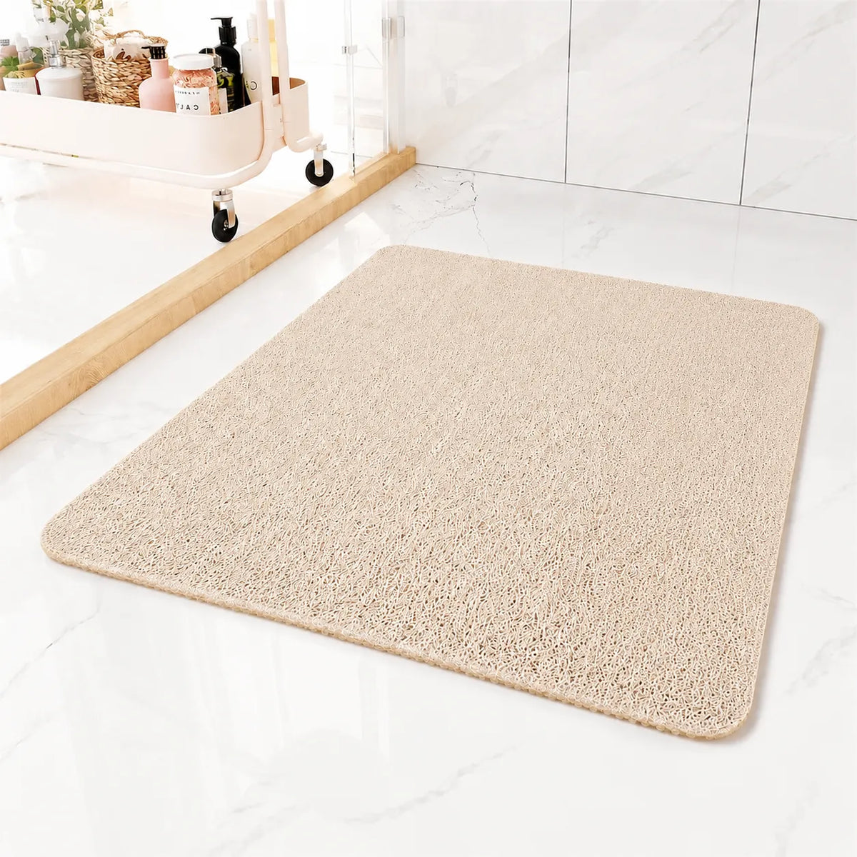 A rectangular beige bath mat with rounded corners, shown on a white marble bathroom floor next to a glass shower door and a small storage cart.