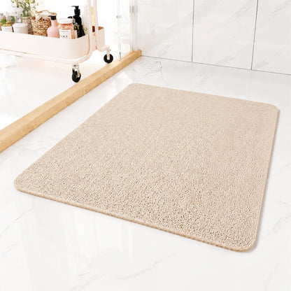 A rectangular beige bath mat with rounded corners, shown on a white marble bathroom floor next to a glass shower door and a small storage cart.