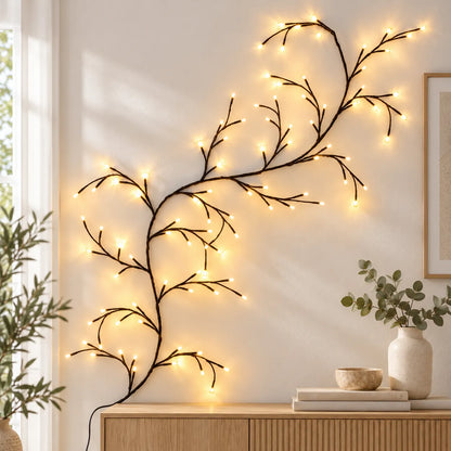 Branch-shaped string wall lights with warm glowing bulbs draped over a light-colored wall above a wooden console table, with a vase and books below and plants at the sides.