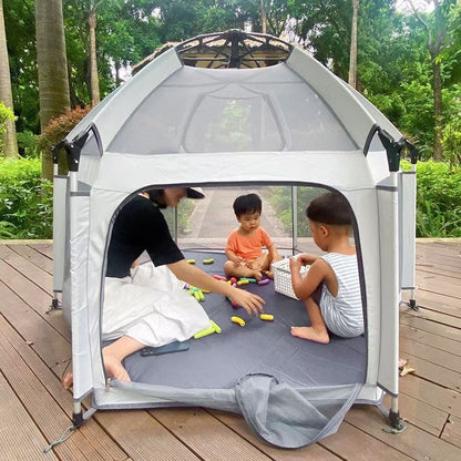Children playing inside a portable playpen on a wooden deck with greenery in the background