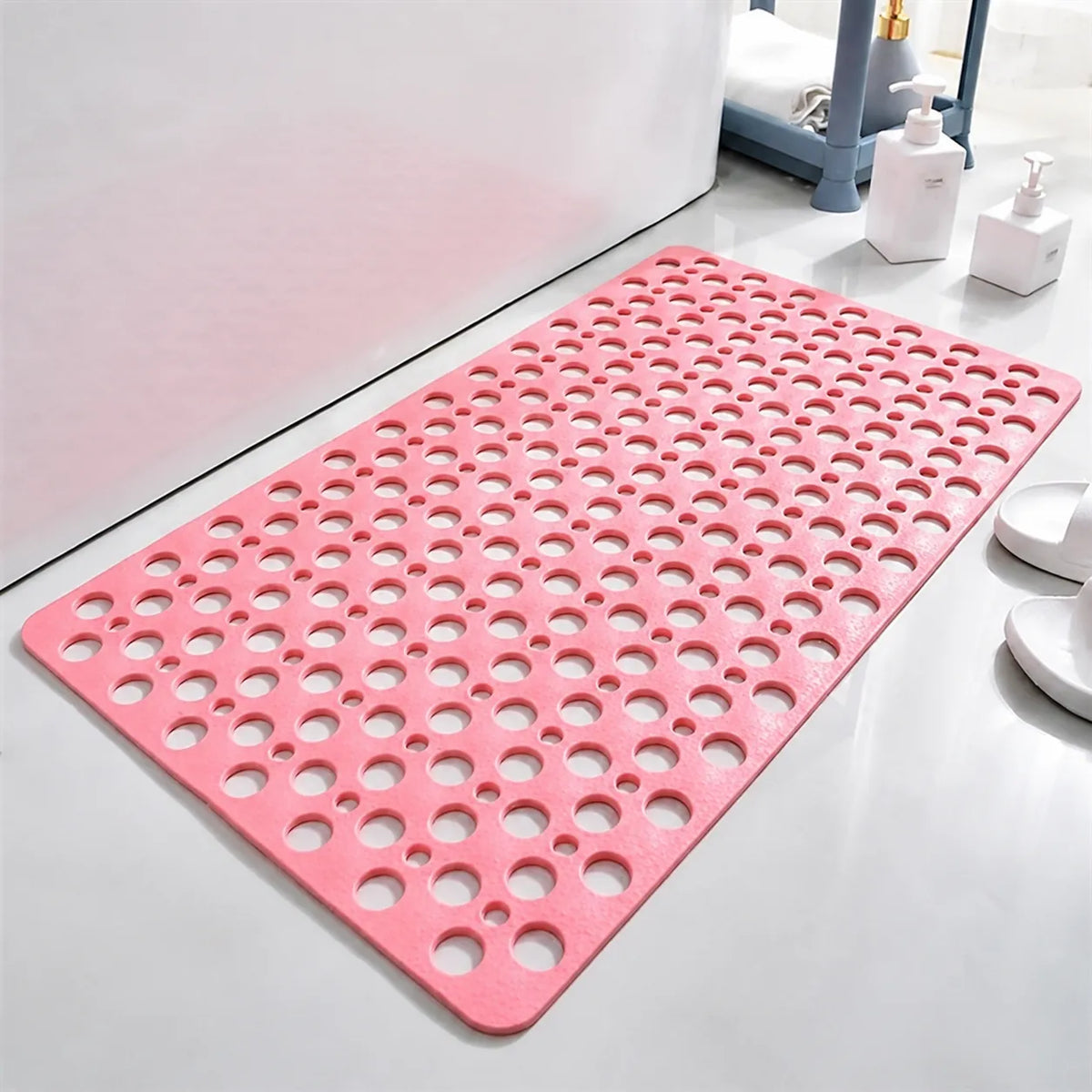 A pink bath mat with a raised circular bubble pattern, shown on a white bathroom floor next to a bathtub, with soap dispensers and a pair of white slippers in the background.