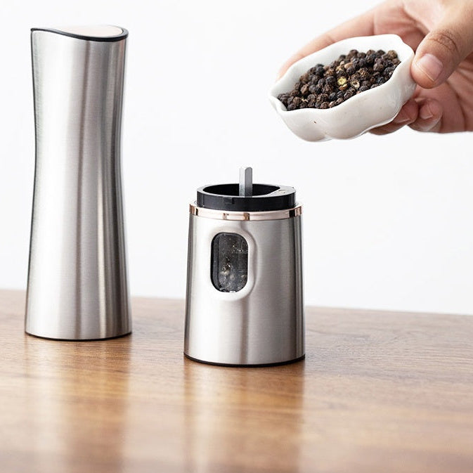 Stainless steel pepper grinder with a small bowl of black peppercorns on a wooden surface.