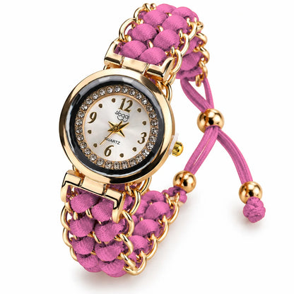 A women's quartz watch with a pink braided cord strap and gold-tone metal links. The round dial is white with gold-tone hands and markers, surrounded by a bezel set with small clear rhinestones.