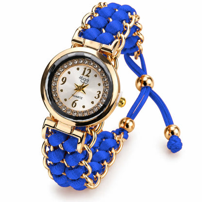 A women's quartz watch with a blue braided cord strap and gold-tone metal links, featuring a round gold-tone case with a crystal-embellished bezel and a white dial.