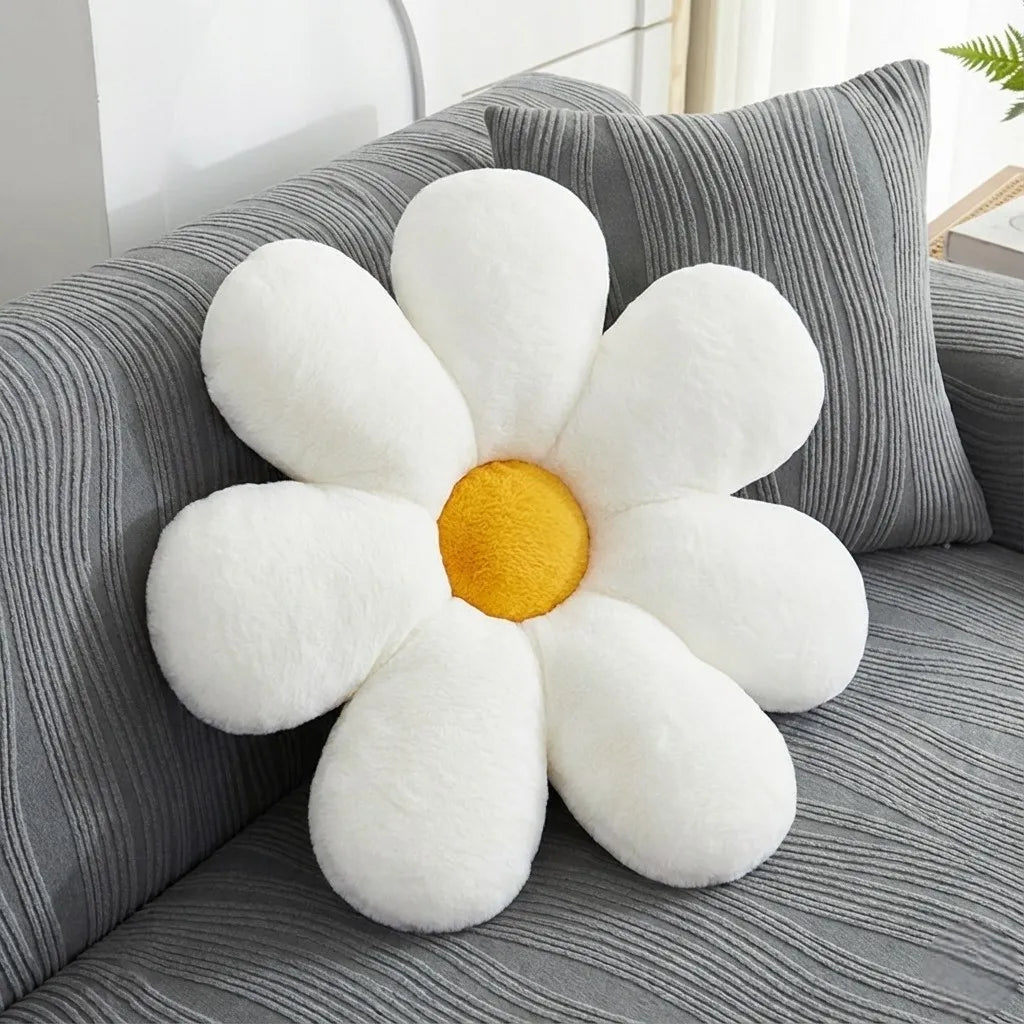 A plush daisy-shaped throw pillow with white petals and a yellow center, resting on a gray striped sofa next to a matching pillow.