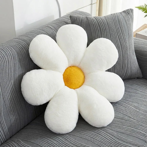 A plush daisy-shaped throw pillow with white petals and a yellow center, resting on a gray striped sofa next to a matching pillow.