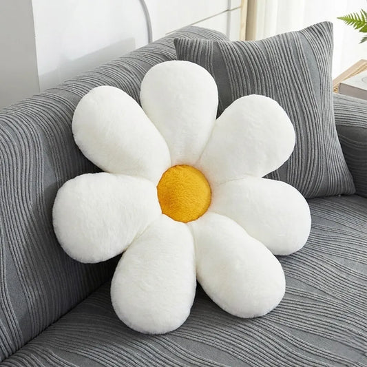 A plush daisy-shaped throw pillow with white petals and a yellow center, resting on a gray striped sofa next to a matching pillow.