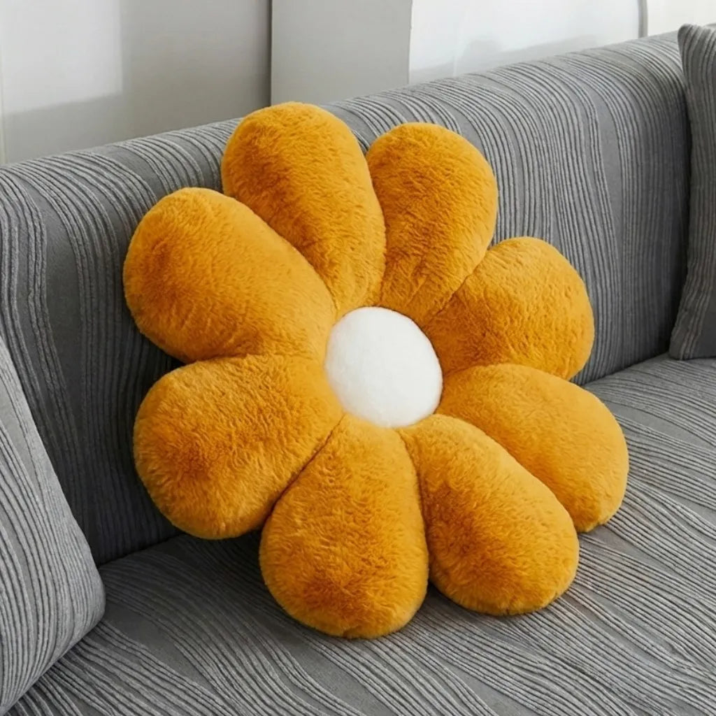 A plush flower-shaped throw pillow with mustard yellow petals and a white center, resting on a gray striped sofa.