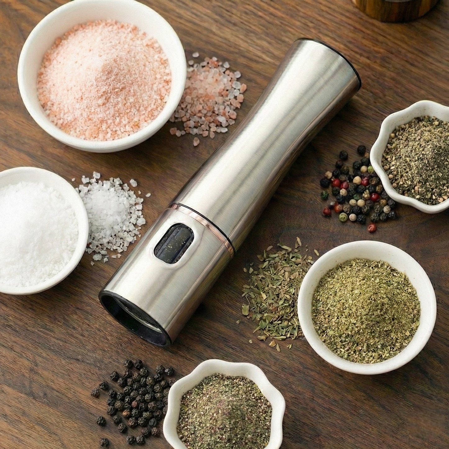 Stainless steel pepper grinder surrounded by various spices on a wooden surface