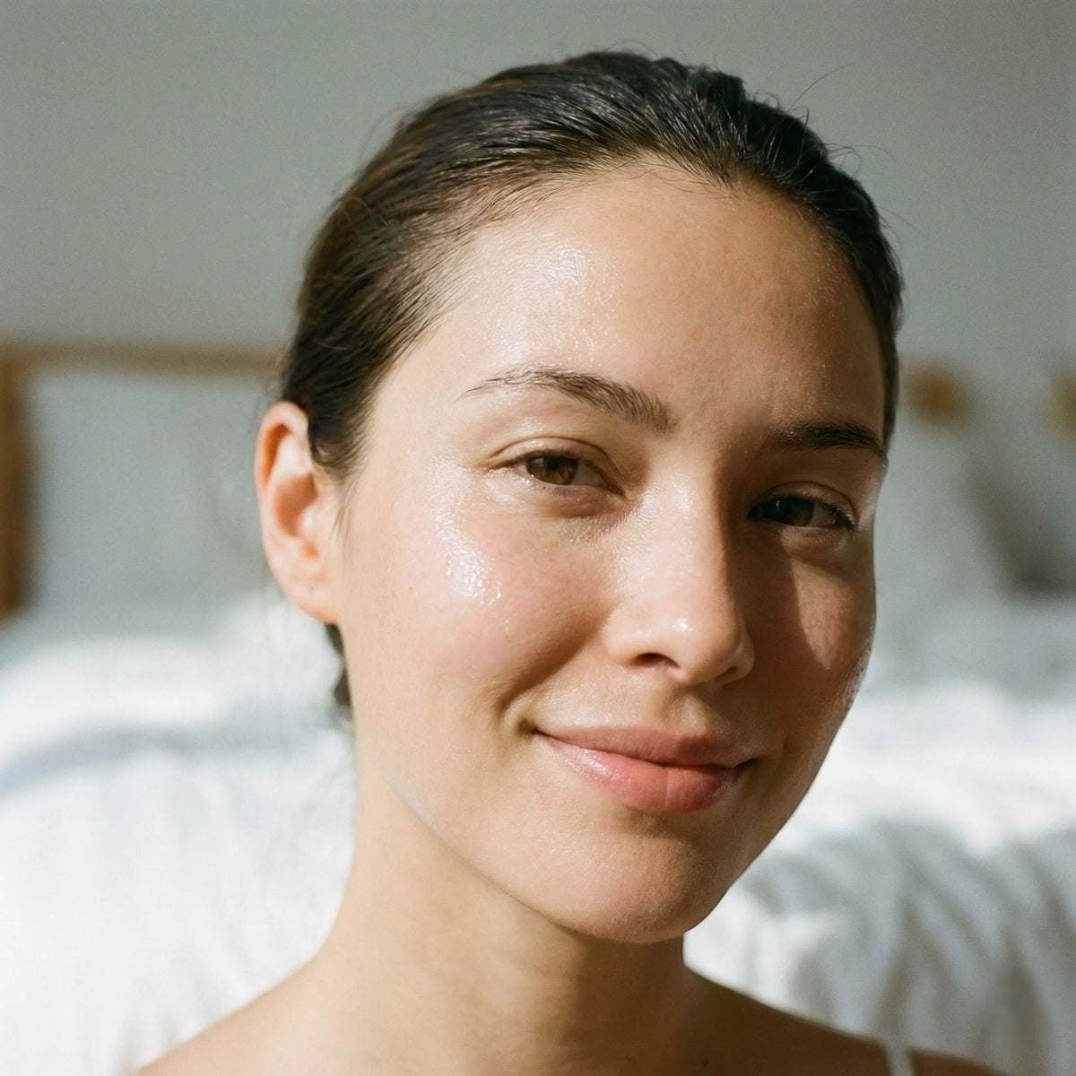 A close-up portrait of a woman with a glossy, hydrated complexion, smiling softly against a softly blurred bedroom background with white bedding.