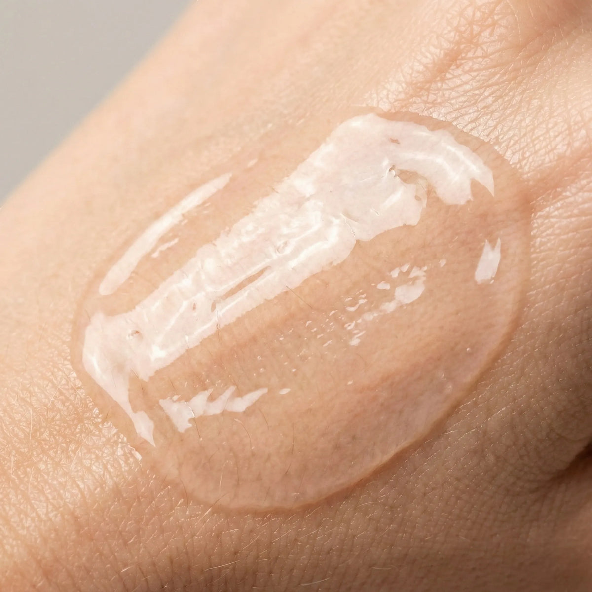 A close-up of a clear, glossy gel or serum swatch applied to human skin, showing its translucent texture and light reflective surface.