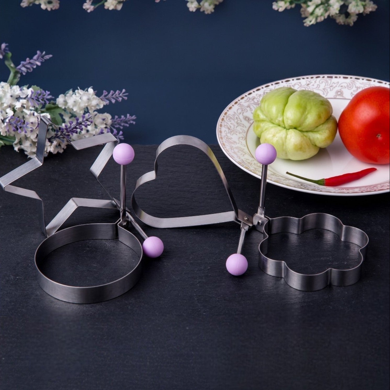 Metallic cookie cutters on a dark surface with a plate of vegetables in the background.