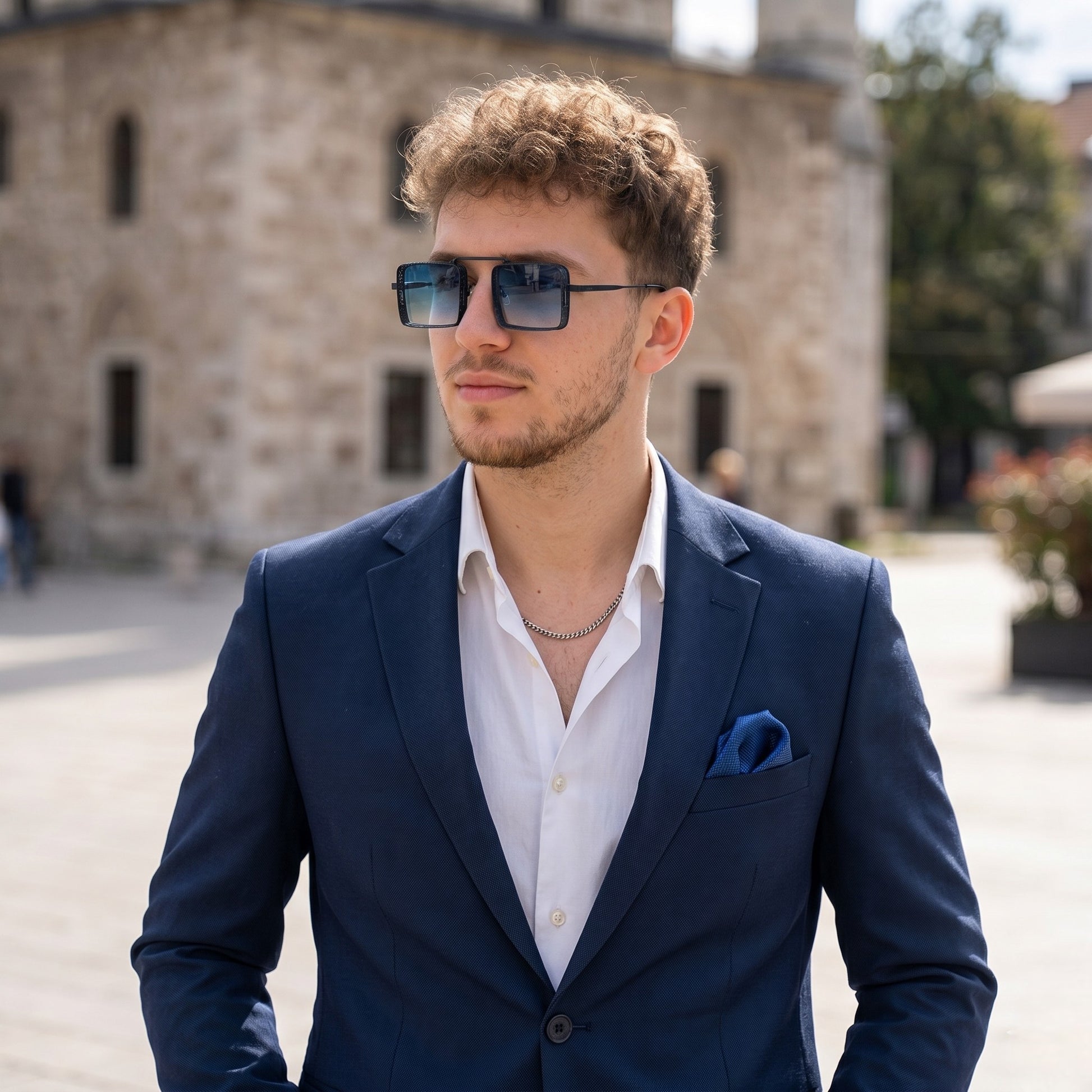 Man wearing a blue suit and sunglasses outdoors with a building in the background