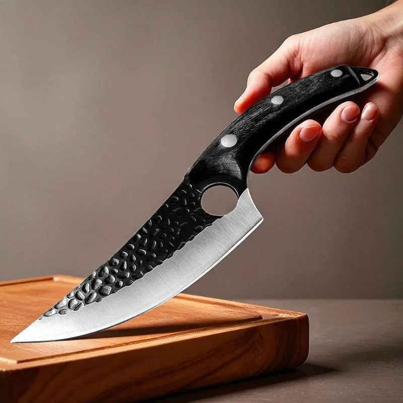 A hand holding a large kitchen knife with a hammered black handle and a silver blade resting on a wooden cutting board against a neutral background.