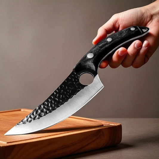 A hand holding a large kitchen knife with a hammered black handle and a silver blade resting on a wooden cutting board against a neutral background.