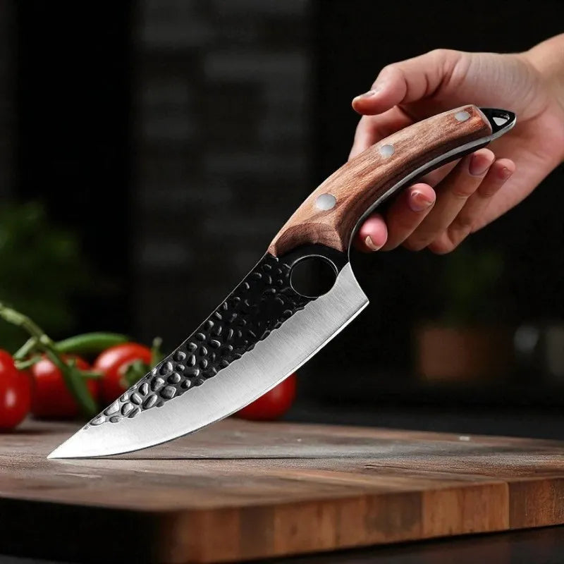 A hand holding a kitchen knife with a hammered black steel blade and a brown wood handle, resting on a wooden cutting board with tomatoes in the background.
