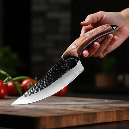 A hand holding a kitchen knife with a hammered black steel blade and a brown wood handle, resting on a wooden cutting board with tomatoes in the background.